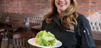 Seattle, Wa  - 142000 - gg - 10/03/2014  -  Caprial Pence is the new chef at the Bookstore Cafe in the Alexis Hotel on First Avenue. After years as an acclaimed chef in Seattle, she moved to Portland. Now she's back in Seattle.  She's holding a couple of new offerings that she has created. Butter lettuce salad with black peper mint dressing and local shaved melon (background), and seared salmon with wild mushrooms and parmeson brado (foreground).